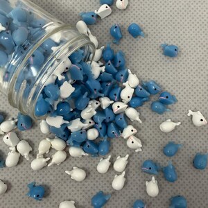 Puede incluir: Un tarro de cristal volcado, derramando una colecci&oacute;n de peque&ntilde;os amuletos con forma de ballena azules y blancas. Los amuletos est&aacute;n esparcidos sobre una superficie gris texturizada, algunos a&uacute;n dentro del tarro. Las ballenas tienen ojos negros y detalles rosas.