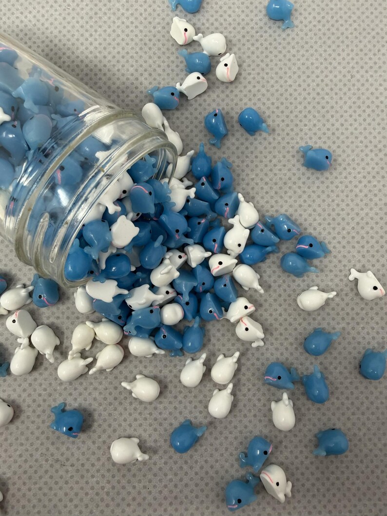 Puede incluir: Un tarro de cristal volcado, derramando una colecci&oacute;n de peque&ntilde;os amuletos con forma de ballena azules y blancas. Los amuletos est&aacute;n esparcidos sobre una superficie gris texturizada, algunos a&uacute;n dentro del tarro. Las ballenas tienen ojos negros y detalles rosas.