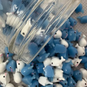 Puede incluir: Una colecci&oacute;n de peque&ntilde;as figuras en forma de ballena, azules y blancas, que se derraman de un tarro de cristal transparente. Las figuras tienen ojos negros y detalles rosas. Las ballenas est&aacute;n esparcidas sobre una superficie gris claro.