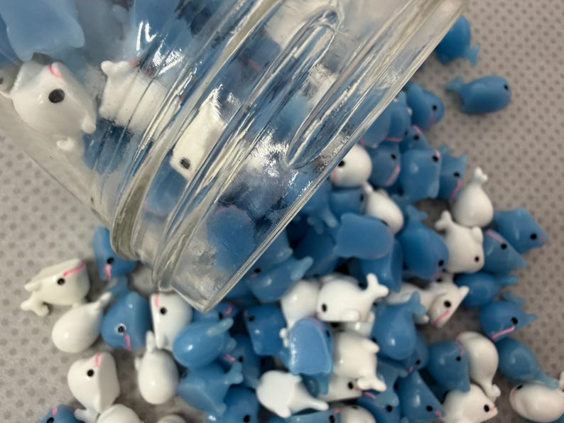 Puede incluir: Una colecci&oacute;n de peque&ntilde;as figuras en forma de ballena, azules y blancas, que se derraman de un tarro de cristal transparente. Las figuras tienen ojos negros y detalles rosas. Las ballenas est&aacute;n esparcidas sobre una superficie gris claro.