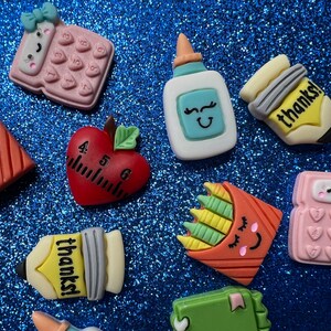 May include: A set of ten colourful, cartoon-style school-themed resin charms. The charms include a red apple with a ruler, a yellow pencil with the word "thanks" on it, a pink calculator, a blue glue bottle, a yellow crayon box, an orange crayon box, a pink calculator with a bow, and a yellow pencil with the word "thanks" on it.