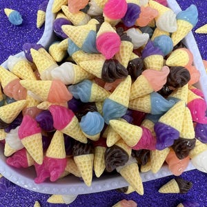 May include: A heart-shaped bowl filled with miniature ice cream cone charms. The cones have yellow waffle bases and colorful scoops of pink, purple, blue, brown, and peach-colored ice cream. Some charms are scattered around the bowl on a purple glitter surface.