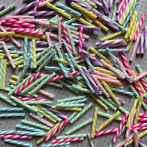 May include: A collection of colorful striped candy sticks in various pastel colors, including pink, yellow, blue, green, and purple. The candy sticks are arranged in a random pattern on a gray surface.