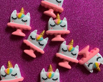 May include: Eight white unicorn cake charms with pink frosting and gold horns. The charms are on a pink glitter background.