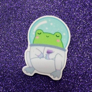 May include: A whimsical sticker featuring a green frog in a space suit with a clear helmet. The frog is smiling with closed eyes, and the suit has purple accents. The background is a sparkling purple.