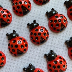 May include: A collection of red and black ladybug-shaped embellishments with black dots. The ladybugs are made of a shiny, glossy material.