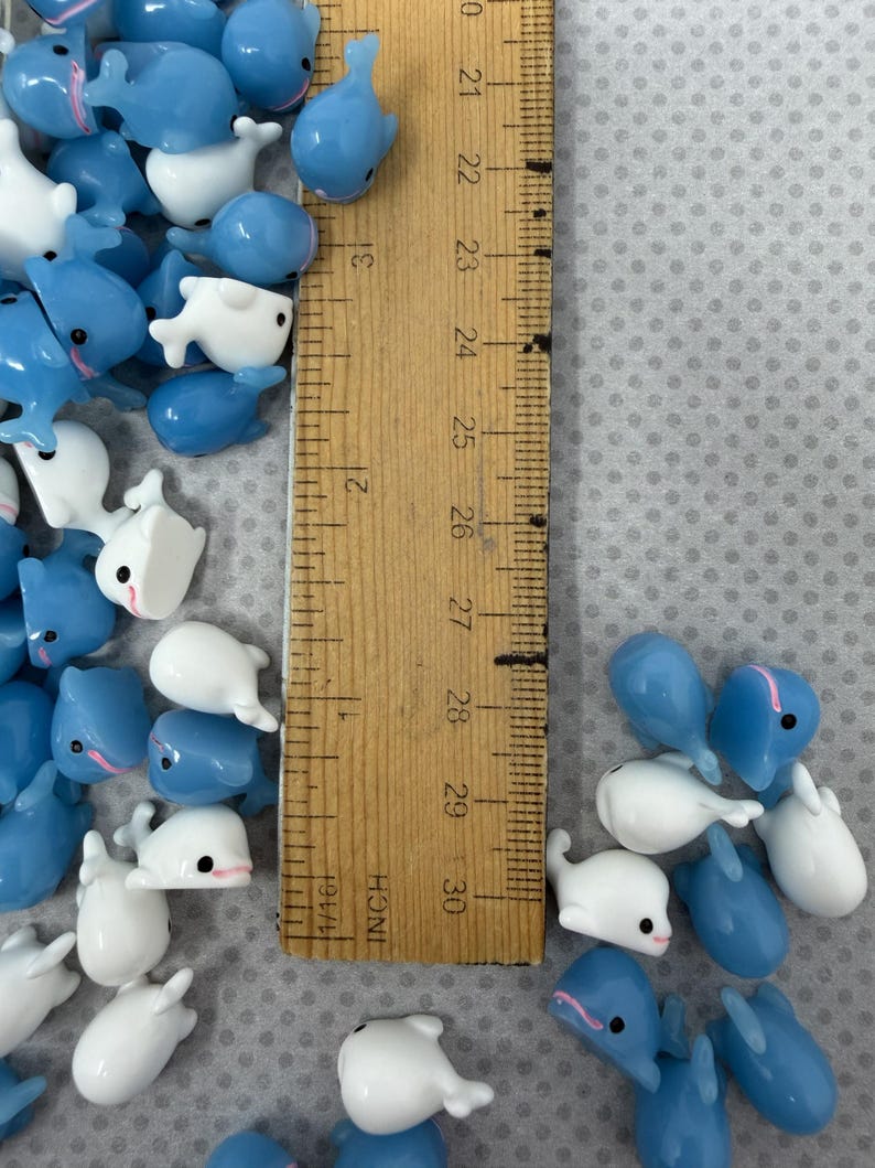 Puede incluir: Una colecci&oacute;n de peque&ntilde;os dijes con forma de ballena, azules y blancos. Los dijes est&aacute;n dispersos alrededor de una regla de madera marcada en pulgadas, con medidas de hasta 76 cm. Algunas ballenas tienen acentos rosas.
