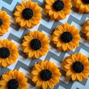 May include: A collection of 12 yellow sunflower-shaped embellishments with black centers. The embellishments are arranged in a pattern on a gray and white chevron background.