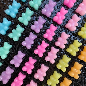 May include: A collection of gummy bear shaped candies in various pastel colors, including pink, blue, green, yellow, and purple. The candies are arranged in rows on a black background.