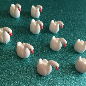 May include: Ten white swan figurines with red beaks on a turquoise glitter background.
