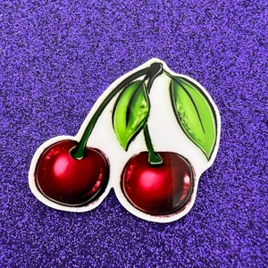 May include: Two red cherries with green leaves on a purple glitter background.