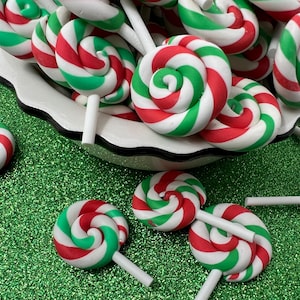 May include: A collection of spiral lollipops in red, green, and white, displayed in a white bowl and scattered on a glittery green surface. The candies have a classic candy cane design.