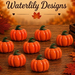 May include: A collection of small, glossy orange ceramic pumpkins with dark green stems. The text Waterlily Designs is displayed at the top, accompanied by a maple leaf icon. The pumpkins are arranged on a wooden surface with scattered autumn leaves.