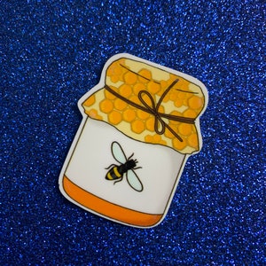 May include: A yellow and white honey jar with a brown ribbon and a bee illustration. The jar is on a blue sparkly background.