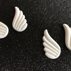 May include: Four white plastic angel wings with a raised design. The wings are on a black sparkly background.