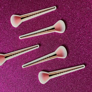 May include: Five pink and gold makeup brushes with fluffy bristles on a sparkly pink background.