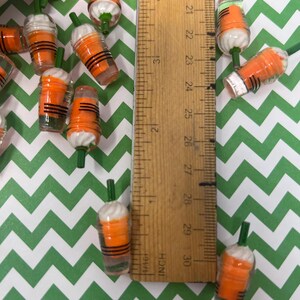 K&ouml;nnte beinhalten: Nahaufnahme einer Gruppe von Miniatur-Bechern aus orangefarbenem und wei&szlig;em Kunststoff mit gr&uuml;nen Strohhalmen. Die Becher sind etwa 2,5 cm hoch und auf einer gr&uuml;n-wei&szlig;en Chevron-Oberfl&auml;che angeordnet. Ein Lineal ist im Hintergrund sichtbar.