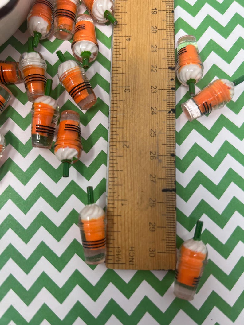 K&ouml;nnte beinhalten: Nahaufnahme einer Gruppe von Miniatur-Bechern aus orangefarbenem und wei&szlig;em Kunststoff mit gr&uuml;nen Strohhalmen. Die Becher sind etwa 2,5 cm hoch und auf einer gr&uuml;n-wei&szlig;en Chevron-Oberfl&auml;che angeordnet. Ein Lineal ist im Hintergrund sichtbar.