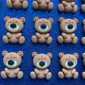 May include: A collection of 12 brown teddy bear-shaped resin embellishments with black eyes and white noses. The bears are sitting with their arms crossed and have a stitched seam down the middle of their bodies.