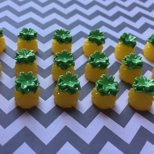 May include: A collection of 18 small, yellow pineapple-shaped charms with green tops. The charms are arranged in rows on a gray and white chevron patterned surface.