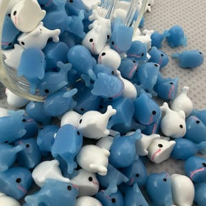 Puede incluir: Un conjunto de peque&ntilde;os dijes en forma de ballena, azules y blancos, con ojos negros y detalles rosas, saliendo de un recipiente de vidrio. Estas figuras de ballena en miniatura son ideales para manualidades o joyer&iacute;a.