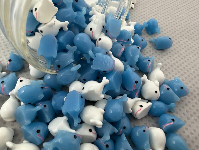Puede incluir: Un conjunto de peque&ntilde;os dijes en forma de ballena, azules y blancos, con ojos negros y detalles rosas, saliendo de un recipiente de vidrio. Estas figuras de ballena en miniatura son ideales para manualidades o joyer&iacute;a.