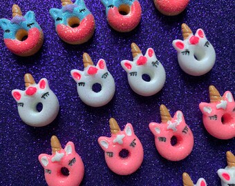 May include: A collection of unicorn-shaped doughnuts with pink, white and blue frosting. The doughnuts have a hole in the centre and are decorated with black eyes and a gold horn.