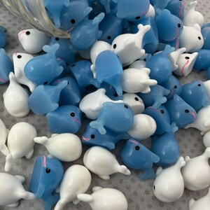 Puede incluir: Un conjunto de peque&ntilde;as figuras de ballenas en tonos azules y blancos. Las figuras tienen un acabado brillante y presentan peque&ntilde;os ojos negros y detalles rosas. Las ballenas est&aacute;n dispersas, lo que sugiere un suministro de manualidades o un art&iacute;culo decorativo.