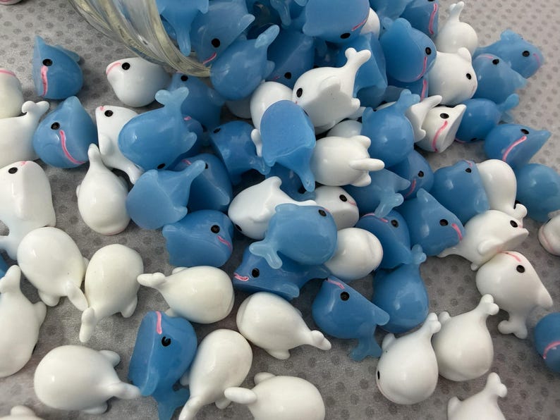 Puede incluir: Un conjunto de peque&ntilde;as figuras de ballenas en tonos azules y blancos. Las figuras tienen un acabado brillante y presentan peque&ntilde;os ojos negros y detalles rosas. Las ballenas est&aacute;n dispersas, lo que sugiere un suministro de manualidades o un art&iacute;culo decorativo.