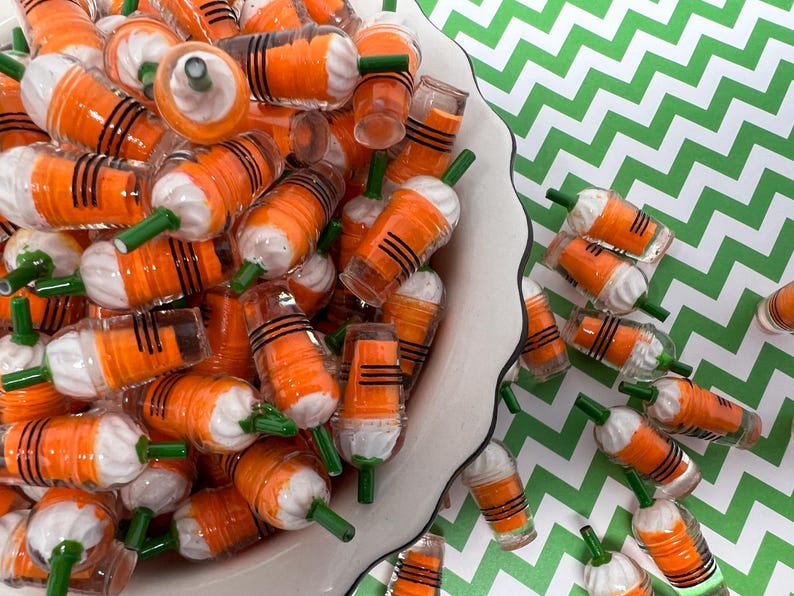 K&ouml;nnte beinhalten: Eine wei&szlig;e Sch&uuml;ssel gef&uuml;llt mit orangefarbenen und wei&szlig;en Miniatur-Plastikbechern mit gr&uuml;nen Strohhalmen. Die Becher haben schwarze Streifen und &auml;hneln Miniatur-Eiskaffee-Getr&auml;nken.