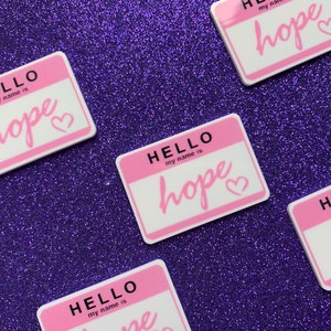 May include: Four pink name tags with the text "HELLO my name is hope" and a heart symbol. The name tags are on a purple sparkly background.