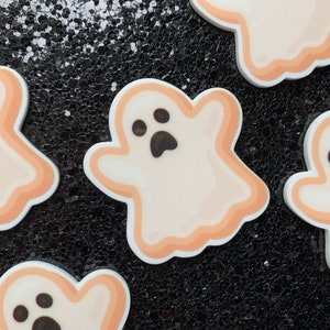 May include: Five white ghost-shaped cookies with black eyes and mouths. The cookies are decorated with a light orange outline.
