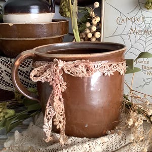 May include: A brown ceramic mug adorned with a delicate, light-colored lace ribbon tied in a bow around its exterior. The mug features a smooth, glazed finish and a single handle, positioned near a book titled Captive Memories by James Terry White.