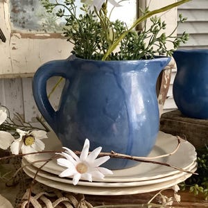 May include: A glossy blue ceramic pitcher with a curved handle sits atop a stack of three white scalloped plates. The pitcher is filled with artificial white daisies and green foliage. A smaller blue creamer pitcher is positioned to the right.