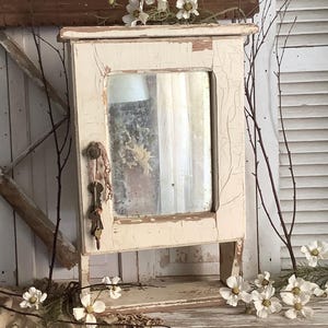 May include: A rectangular wall-mounted cabinet with a distressed off-white painted finish and a central mirror panel. A small round knob on the door holds a cluster of three metal keys on a string. The cabinet features visible cracks in the paint.