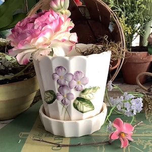 May include: A white ceramic flower pot with a matching saucer, decorated with purple violet flowers and green leaves. The pot is filled with pink and white flowers. A rustic, vintage-style decorative piece.