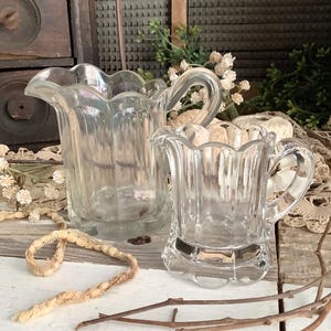 May include: Two clear glass cream pitchers with scalloped rims and handles. The pitchers are different sizes and sit on a white wooden surface. Decorative elements include dried flowers and branches.