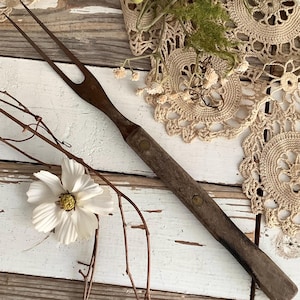 May include: An antique carving fork with a dark wooden handle and two tines. The handle is riveted with brass pins. The fork is resting on a distressed white wooden surface, with floral and lace accents.