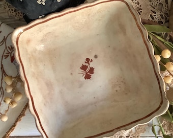 May include: A square ceramic bowl with a cream-colored base, featuring a scalloped rim outlined in a thin brown line. A small, singular brown botanical leaf motif is centered on the interior floor of the bowl, which shows light brown staining.