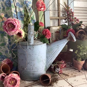 May include: A distressed, pale blue metal watering can overflowing with pink and red blooms. Small terracotta pots and more flowers are arranged around the can. The scene is set against a backdrop of a window and floral fabric, creating a rustic, garden aesthetic.