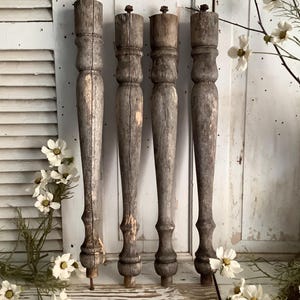 May include: Four tall, weathered wooden furniture legs with a cylindrical, turned design. Each leg features a dark, distressed gray-brown finish with visible wood grain, vertical cracks, and a small metal bolt protruding from the top end.