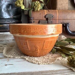 May include: A glazed orange ceramic mixing bowl featuring a relief pattern of a house with windows and a figure standing outside. The rim is decorated with a repeating geometric motif. The bowl has a wide opening and a tapered base.