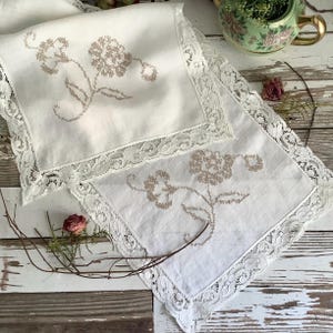 May include: Two white linen handkerchiefs with embroidered floral designs in tan. Each handkerchief has a delicate lace trim around the edges. The items are placed on a rustic, weathered wooden surface.