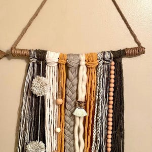 May include: Handmade wall hanging featuring a variety of textures and colors. The piece includes strands of yarn in white, black, gray, and mustard yellow, accented with wooden beads and pom-poms. A decorative item for home decor.