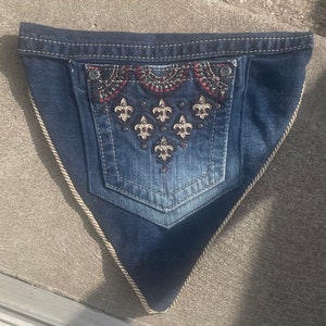 May include: A dark blue denim triangle bandana with a decorative pocket featuring a fleur-de-lis design with gold and red embellishments.