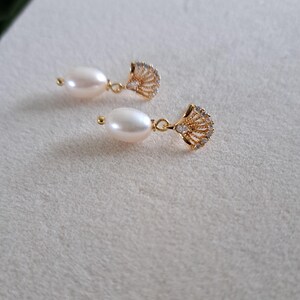 May include: A pair of elegant drop earrings featuring lustrous, oval-shaped pearls suspended from gold-colored, fan-shaped pendants. The pendants are embellished with small, sparkling crystals, adding a touch of glamour to the jewelry.