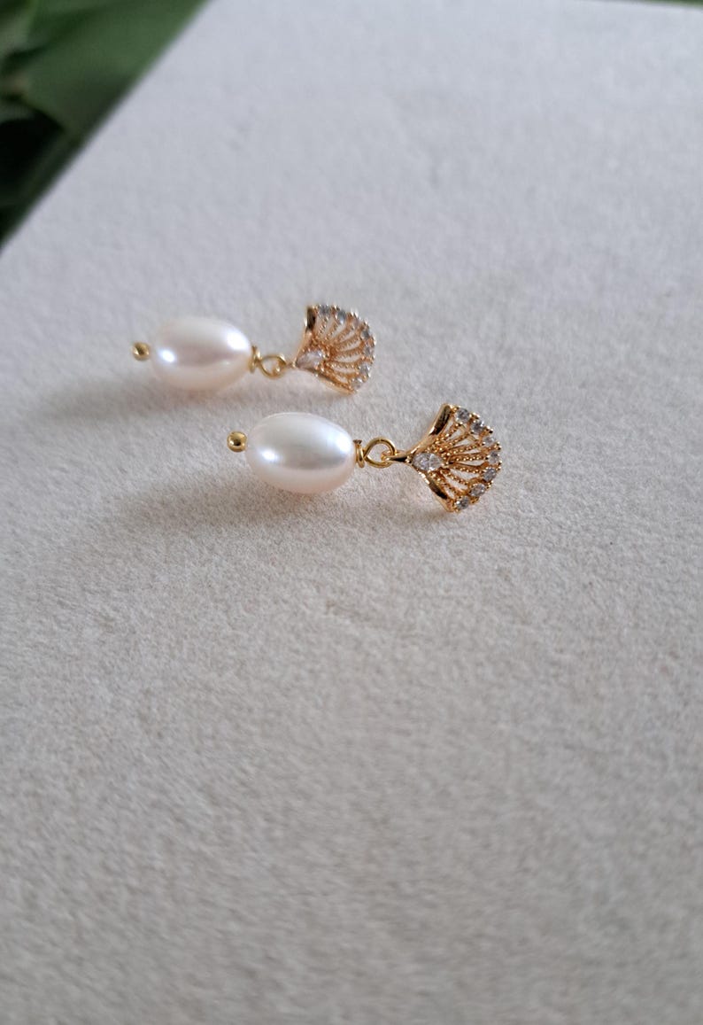 May include: A pair of elegant drop earrings featuring lustrous, oval-shaped pearls suspended from gold-colored, fan-shaped pendants. The pendants are embellished with small, sparkling crystals, adding a touch of glamour to the jewelry.