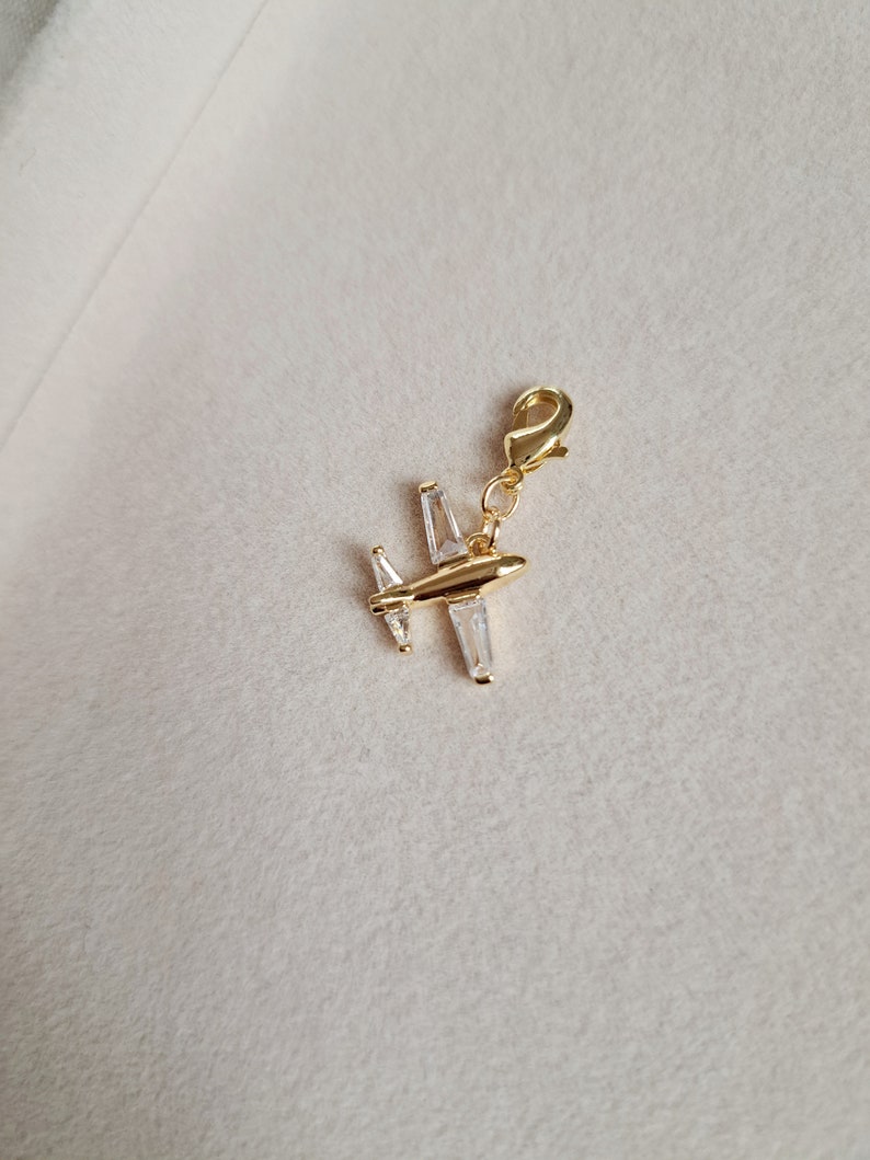 May include: Gold airplane charm with clear crystal accents. The charm has a lobster clasp for attaching to a bracelet or necklace.