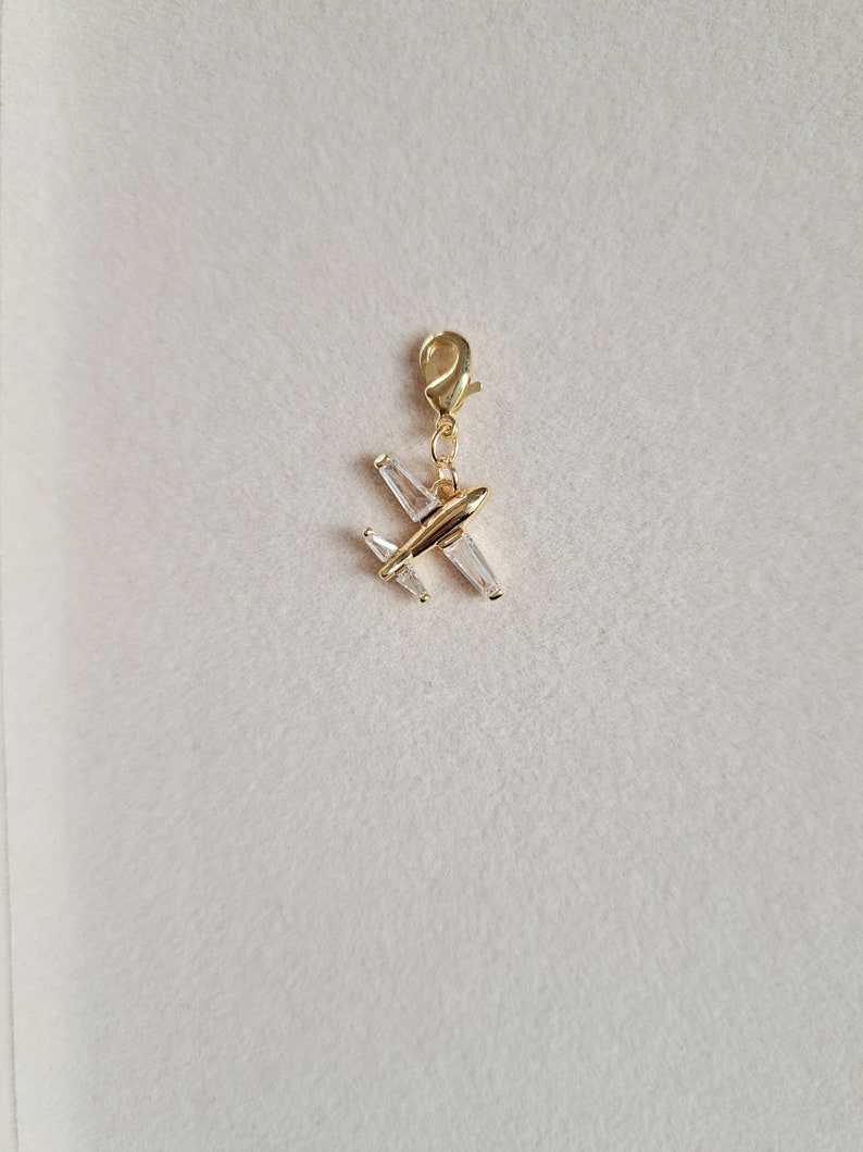 May include: Gold airplane charm with clear crystal accents. The charm has a lobster clasp for attaching to a bracelet or necklace.