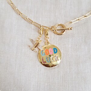 May include: Gold chain necklace with a toggle clasp and two charms. One charm is a small airplane and the other is a round charm with the words "GOOD LUCK" in colorful enamel letters.